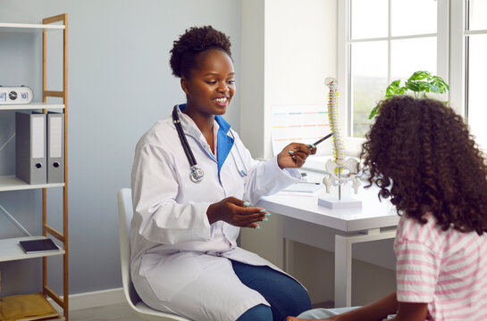 Friendly dark-skinned female doctor demonstrates about possible back problems and prevention of spinal curvature. Girl listens to the doctor, looking at the anatomical model of the spine in the office