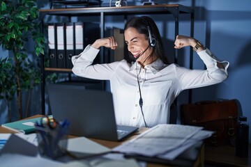 Young brunette woman wearing call center agent headset working late at night showing arms muscles...