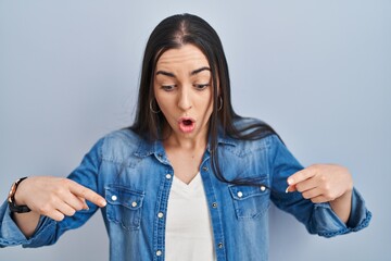 Hispanic woman standing over blue background pointing down with fingers showing advertisement, surprised face and open mouth
