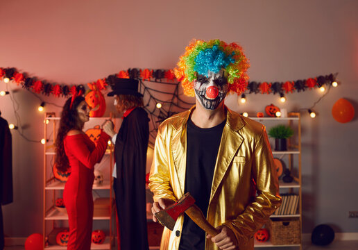 Portrait of scary man clown in costume with axe chopper on Halloween celebration indoors. Smiling crazy male comic have fun on all saints hollows eve party at home. Masquerade, festive concept.