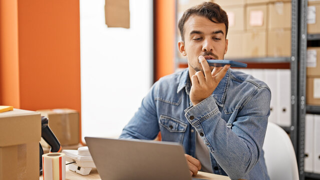 Young hispanic man ecommerce business worker sending voice message by smartphone using laptop at office
