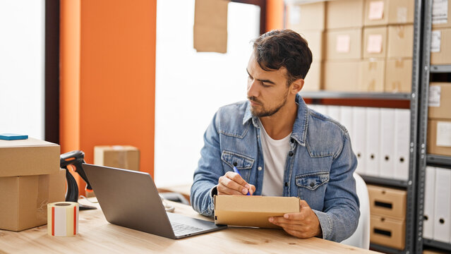 Young hispanic man ecommerce business worker using laptop writing on package at office