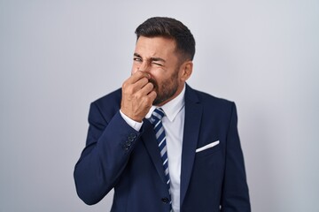Handsome hispanic man wearing suit and tie smelling something stinky and disgusting, intolerable...