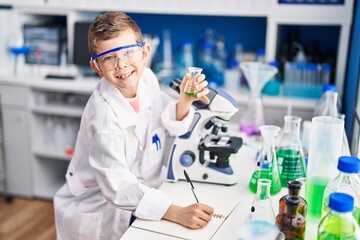 Blond child wearing scientist uniform writing on notebook at laboratory