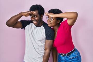 Obraz premium Young african american couple standing over pink background very happy and smiling looking far away with hand over head. searching concept.