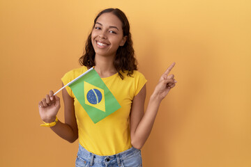 Young hispanic woman holding brazil flag with a big smile on face, pointing with hand finger to the...