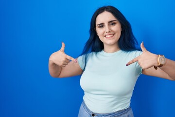 Fototapeta premium Young modern girl with blue hair standing over blue background looking confident with smile on face, pointing oneself with fingers proud and happy.