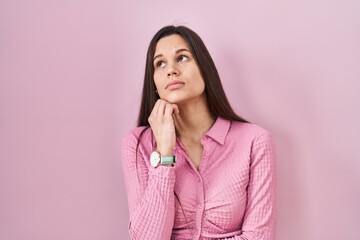 Fototapeta premium Young hispanic woman standing over pink background with hand on chin thinking about question, pensive expression. smiling with thoughtful face. doubt concept.