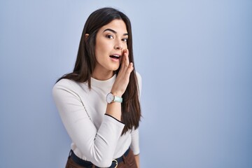 Young brunette woman standing over blue background hand on mouth telling secret rumor, whispering malicious talk conversation - obrazy, fototapety, plakaty