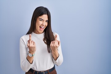 Young brunette woman standing over blue background showing middle finger doing fuck you bad...