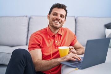 Young hispanic man using laptop and drinking coffee sitting on floor at home