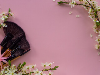 Bouquet of cosmetic brushes tied with a pink ribbon with a bow on pink background with cherry flowers and copy space. Women and Mothers Day gift. Concept of beauty and femininity. Buying gifts online.