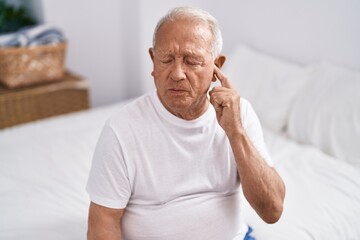 Senior grey-haired man sitting on bed with suffering for ear pain at bedroom - obrazy, fototapety, plakaty