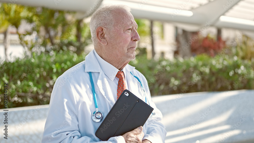 Poster senior grey-haired man doctor standing with serious expression holding touchpad at park