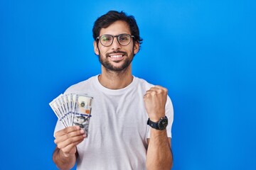 Handsome latin man holding dollars banknotes screaming proud, celebrating victory and success very excited with raised arms