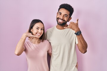 Young hispanic couple together over pink background smiling doing phone gesture with hand and fingers like talking on the telephone. communicating concepts.