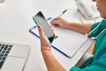 Young beautiful hispanic woman doctor using smartphone writing on document at clinic