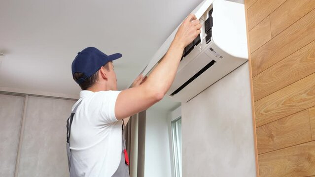 Repairman in work uniform opens air conditioner tor repair. Male worker carefully examines equipment in modern premise. Concept of service at home