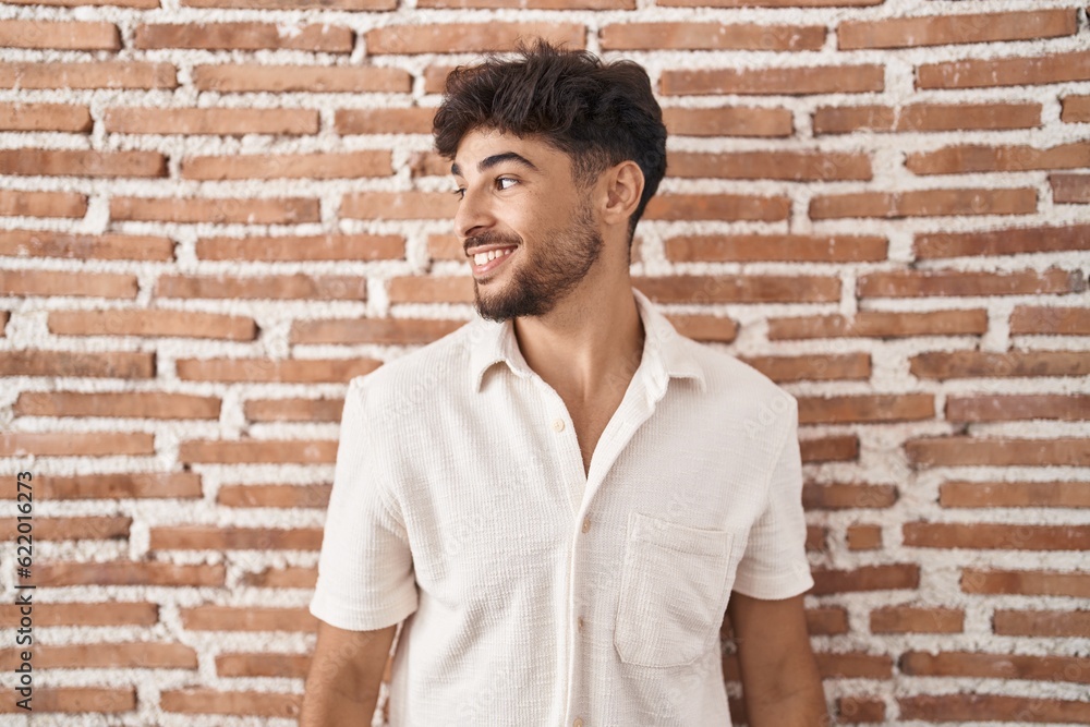 Canvas Prints Arab man with beard standing over bricks wall background looking away to side with smile on face, natural expression. laughing confident.