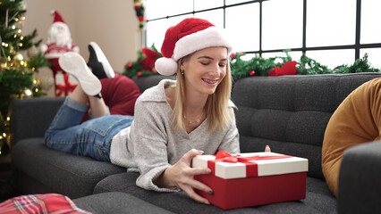 Young blonde woman unpacking gift lying on the sofa by christmas tree at home