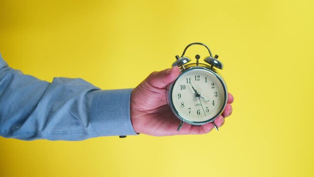 Man's hand hand holds an alarm clock on a yellow background, a man shows the time 15,45 o'clock on an isolated background.