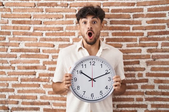 Arab man with beard holding big clock afraid and shocked with surprise and amazed expression, fear and excited face.