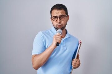 Hispanic man holding reporter microphone and clipboard puffing cheeks with funny face. mouth...