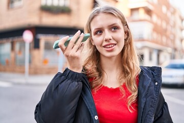 Young blonde woman smiling confident listening audio message by the smartphone at street
