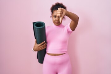 Young hispanic woman with curly hair holding yoga mat over pink background looking unhappy and...