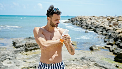 Young hispanic man tourist wearing swimsuit applying sunscreen at the beach