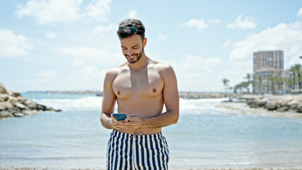 Young hispanic man tourist wearing swimsuit using smartphone smiling at the beach