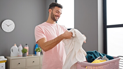 Young hispanic man smiling confident smelling clean towel at laundry room