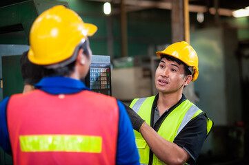 Two young asian male technicians wearing safety uniform workwear, helmets, vest and gloves are Training to work at the lathe control cabinet and inspecting parts intently In industrial plants.