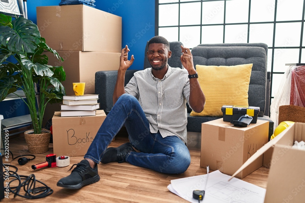 Sticker African american man sitting on the floor at new home gesturing finger crossed smiling with hope and eyes closed. luck and superstitious concept.