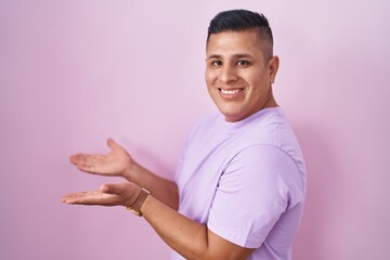 Young hispanic man standing over pink background pointing aside with hands open palms showing copy space, presenting advertisement smiling excited happy