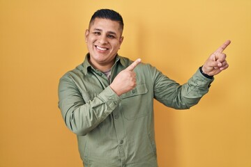 Hispanic young man standing over yellow background smiling and looking at the camera pointing with two hands and fingers to the side.