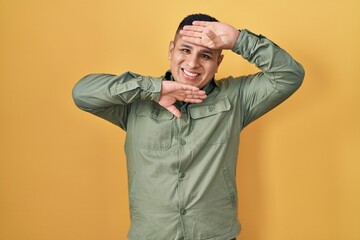 Hispanic young man standing over yellow background smiling cheerful playing peek a boo with hands...