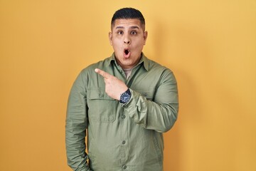 Hispanic young man standing over yellow background surprised pointing with finger to the side, open mouth amazed expression.