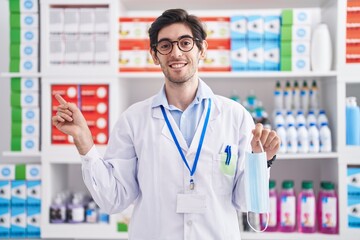 Young hispanic man working at pharmacy drugstore holding safety mask smiling happy pointing with hand and finger to the side