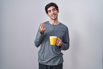 Young hispanic man wearing pajama drinking a cup of coffee doing money gesture with hands, asking...