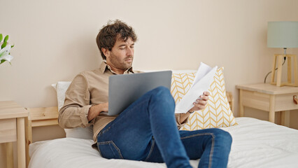 Young hispanic man using laptop reading document at bedroom