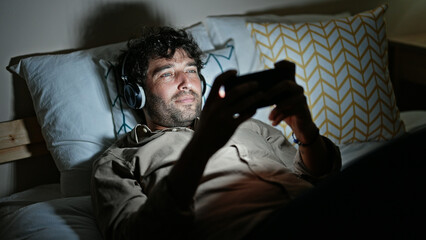 Young hispanic man watching video on smartphone lying on bed at bedroom