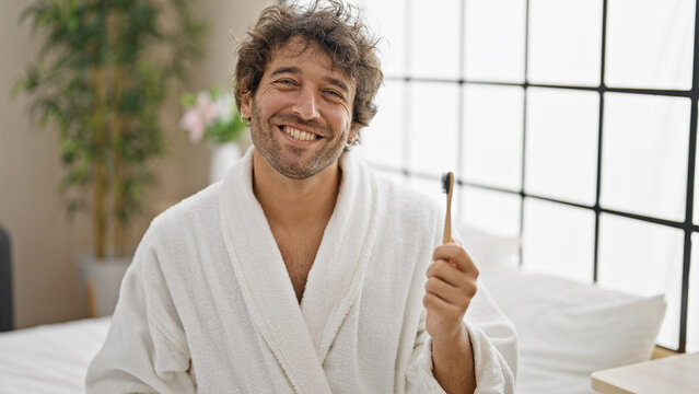 Young hispanic man wearing bathrobe holding wooden toothbrush smiling at bedroom