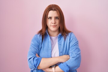 Fototapeta premium Young hispanic woman with red hair standing over pink background skeptic and nervous, disapproving expression on face with crossed arms. negative person.