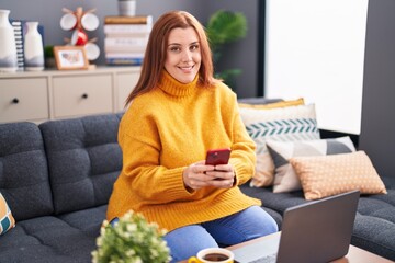 Young beautiful plus size woman using smartphone and laptop sitting on sofa at home