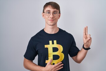 Caucasian blond man wearing bitcoin t shirt smiling swearing with hand on chest and fingers up,...