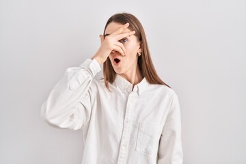 Young caucasian woman standing over isolated background peeking in shock covering face and eyes with hand, looking through fingers with embarrassed expression.