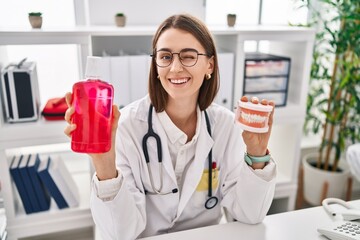Young caucasian dentist woman holding denture and mouthwash winking looking at the camera with sexy...