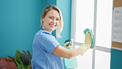 Young blonde woman professional cleaner cleaning window smiling at the office