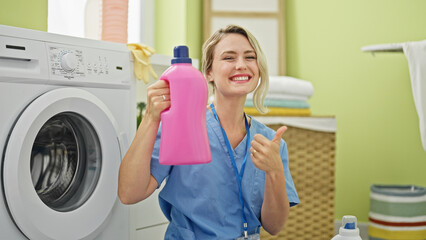 Young blonde woman professional cleaner holding detergent bottle doing thumb up gesture at laundry room